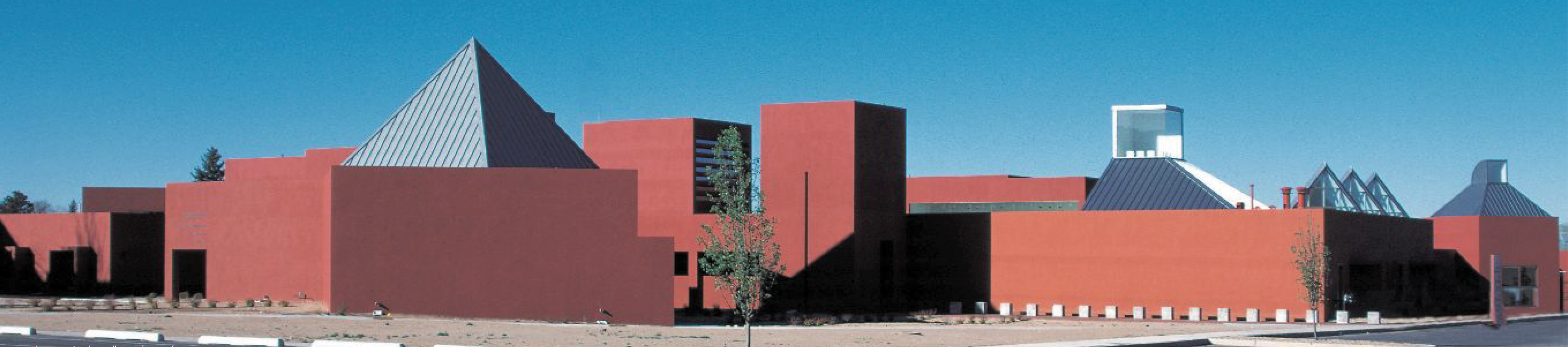 Exterior view of the Visual Arts Center buildings at the Santa Fe Midtown Site, featuring bold red stucco walls and distinctive geometric roof forms designed by architect Ricardo Legorreta, set against a clear blue sky.