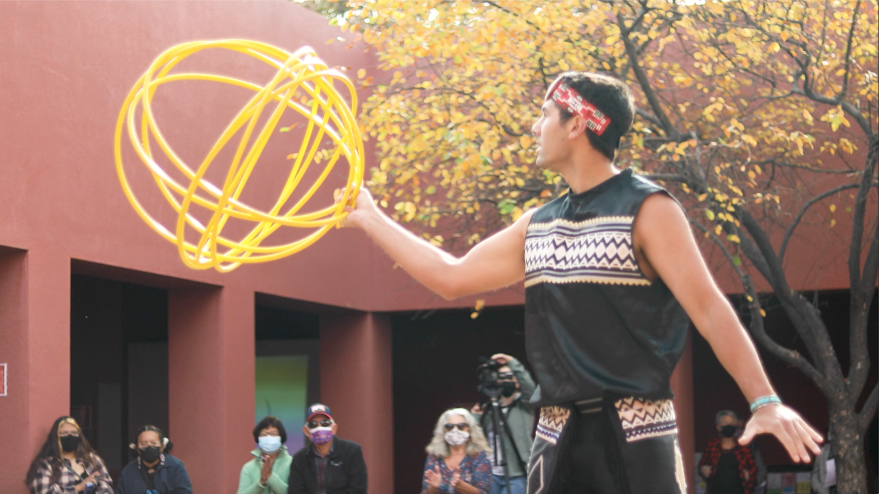 A Native American hoop dancer performs outdoors, skillfully spinning bright yellow hoops. The dancer wears traditional attire with geometric patterns and a red headband, while an audience, many wearing masks, watches against the backdrop of the SFAI buildings.